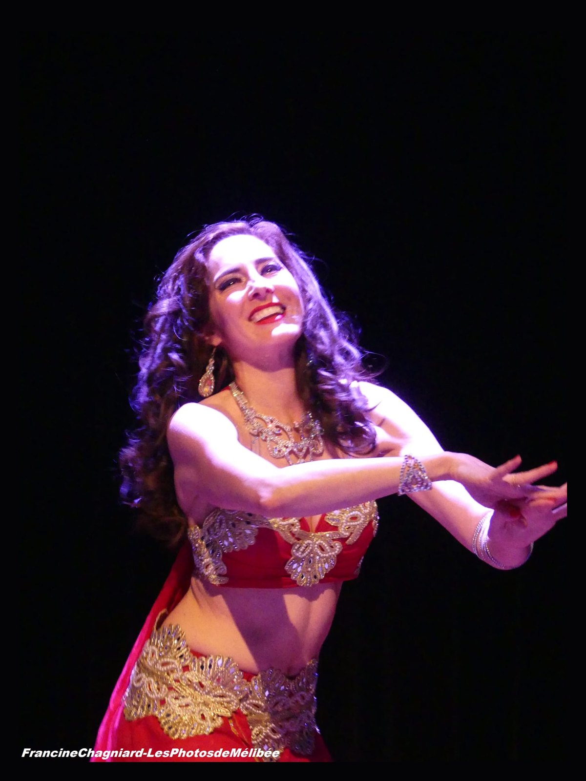 Ciya en costume rouge et or, souriante, cheveux au vent sur scène
